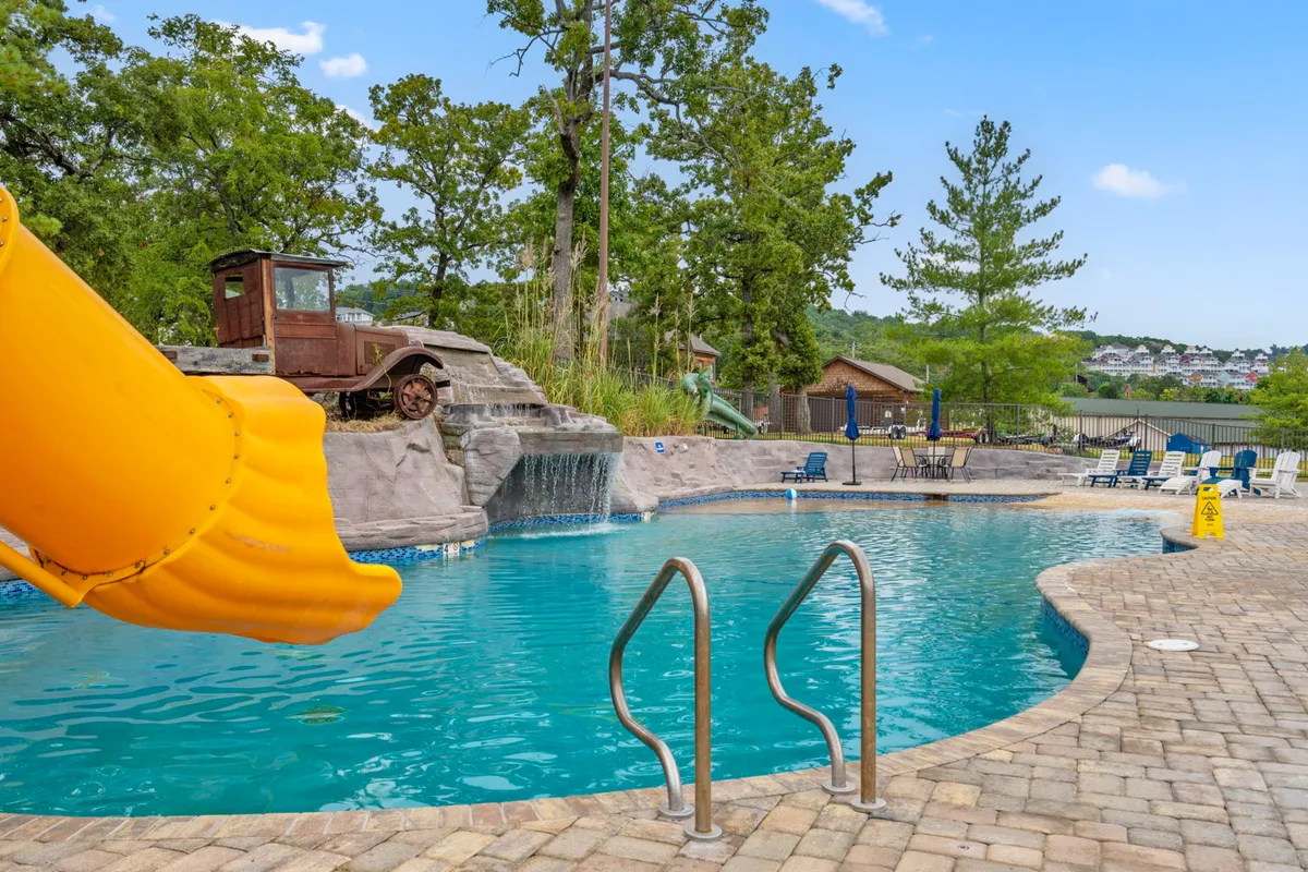 A vibrant scene of the resort’s outdoor pool area, featuring a sparkling pool with a water slide curving into the water, an eye-catching rock formation, a vintage old truck, and inviting lounge chairs scattered around the edge. The lively atmosphere is perfect for summer relaxation and family fun.