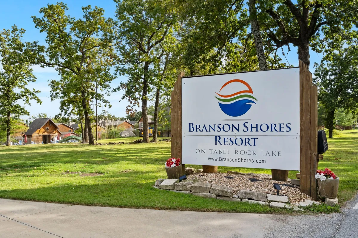 A welcoming exterior shot of the Branson Shores Resort sign, surrounded by leafy trees and cabins nearby. The image sets a friendly, inviting tone and subtly situates the property within the local landscape, hinting at easy access to both nature and attractions.