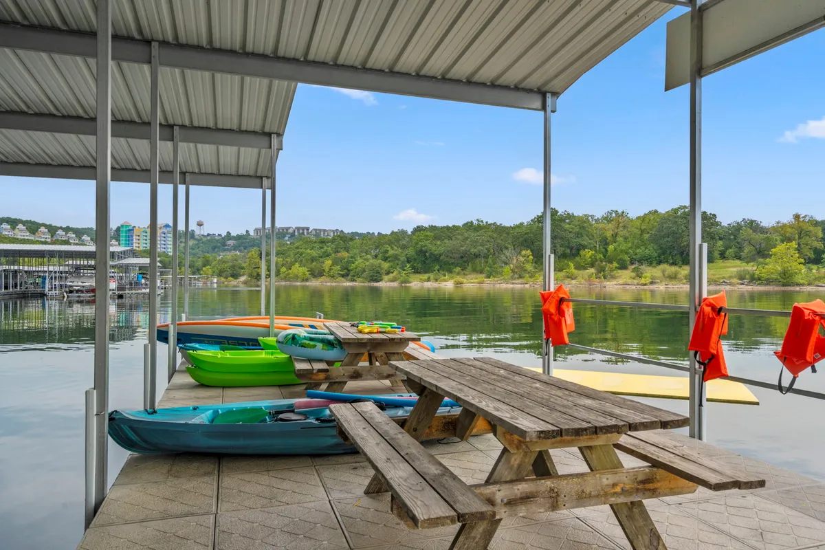 Kayaks rest on the shoreline next to a picnic table, while life jackets are ready for use. The clear water and surrounding greenery create a sense of peaceful adventure. This setting invites guests to kayak, picnic, fish, or simply relax by the water, with the lake itself as the central focus.