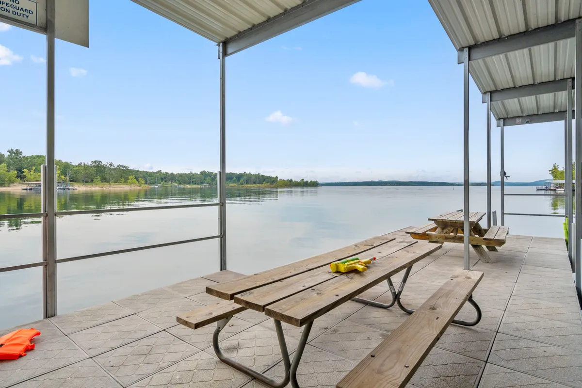 Several picnic tables are positioned beside the calm waters of the lake, with a distant shoreline visible across the water. The scene is tranquil, emphasizing the natural beauty and the opportunities for lakeside picnics, fishing, and relaxation.