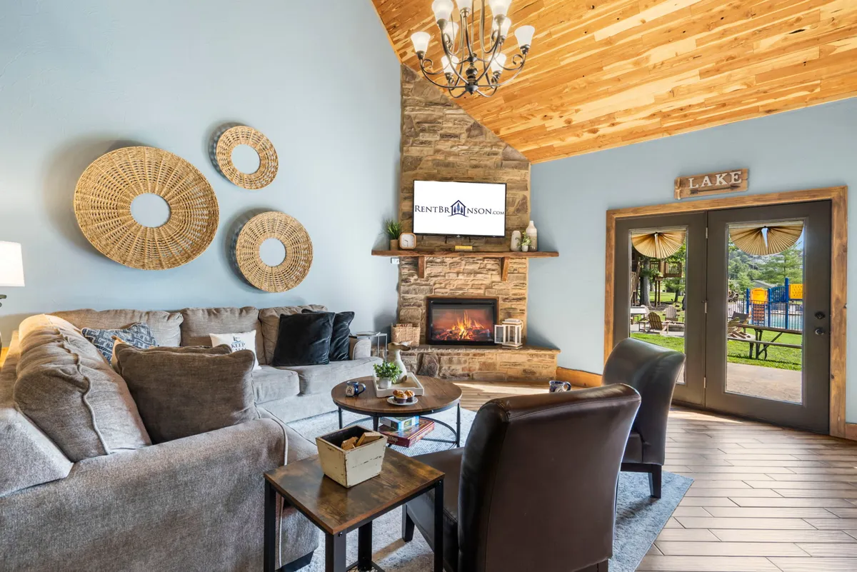 This image features a comfortable sectional sofa positioned in front of a stone fireplace, with decorative wall art and a flat-screen TV. Double doors lead outdoors, and the space is bathed in natural light, highlighting the warm wood floors and inviting atmosphere.