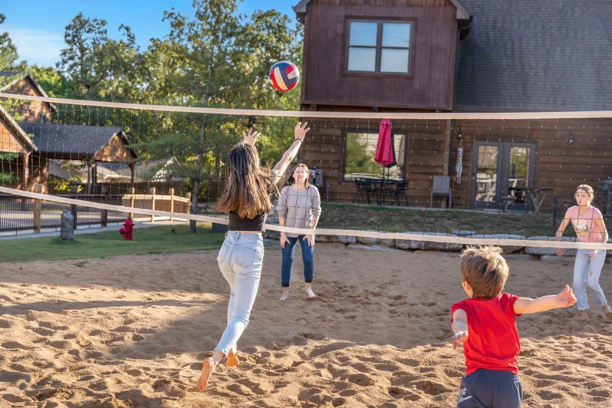An energetic match of volleyball underway on the sand volleyball court, with participants leaping and stretching for the ball. A charming house is visible in the background, anchoring the scene in the resort environment. The image exudes playful group activity and outdoor enjoyment.