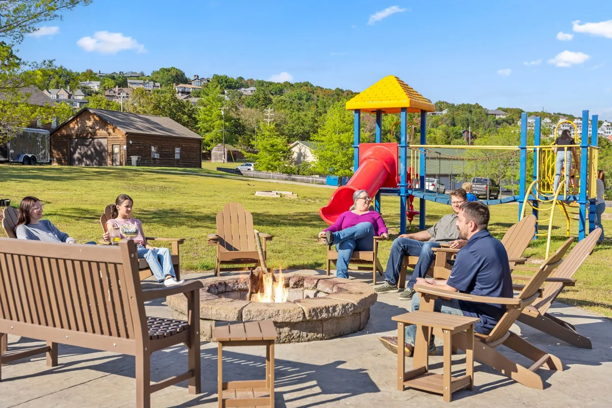 A communal outdoor area with a fire pit, surrounded by chairs and a playground in the background. Guests are seated around the fire, sharing stories, while children play on the playground as the sun sets.