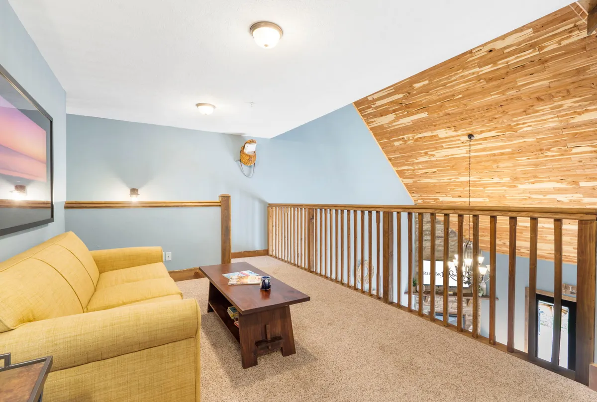 A loft sitting area features a yellow sofa, a wooden coffee table, and a wooden railing overlooking the main living area. Blue walls create a cozy, intimate nook, ideal for quiet moments with coffee or reading.