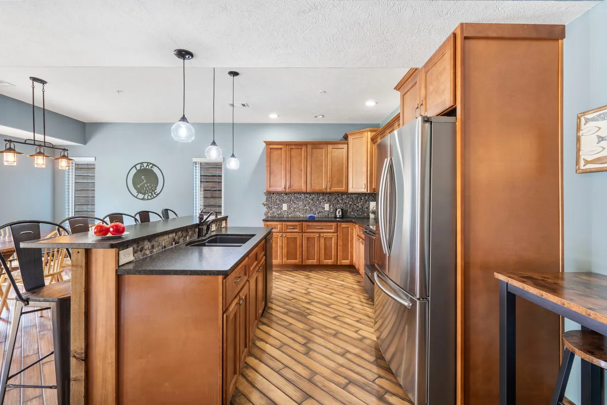 The photograph showcases a modern kitchen with a prominent kitchen island, stainless steel appliances, and wooden cabinetry. Pendant lighting above the island creates a welcoming, functional workspace perfect for shared cooking experiences.