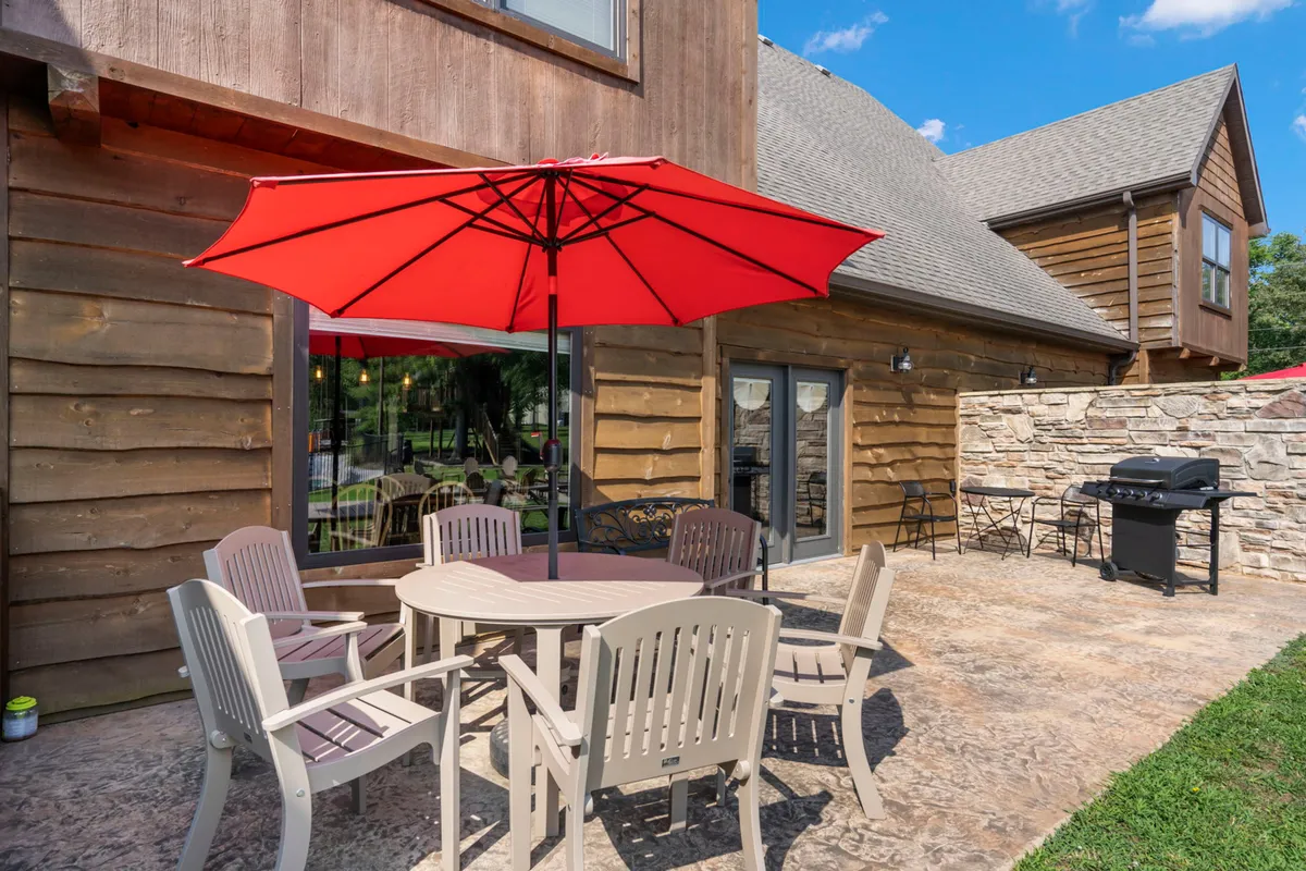 An inviting outdoor dining area set beneath a red umbrella, with a grill and sturdy stone wall nearby. The table is set for a meal, and the space feels bright and cheerful—ideal for both casual breakfasts and evening gatherings.