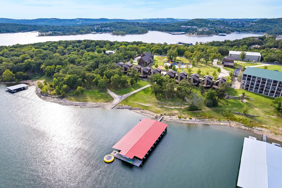 A tranquil lakeside view with a dock extending into the water, cabins nestled by the shoreline, and lush greenery all around. Activities like boating, fishing, and picnicking are suggested by the scene.