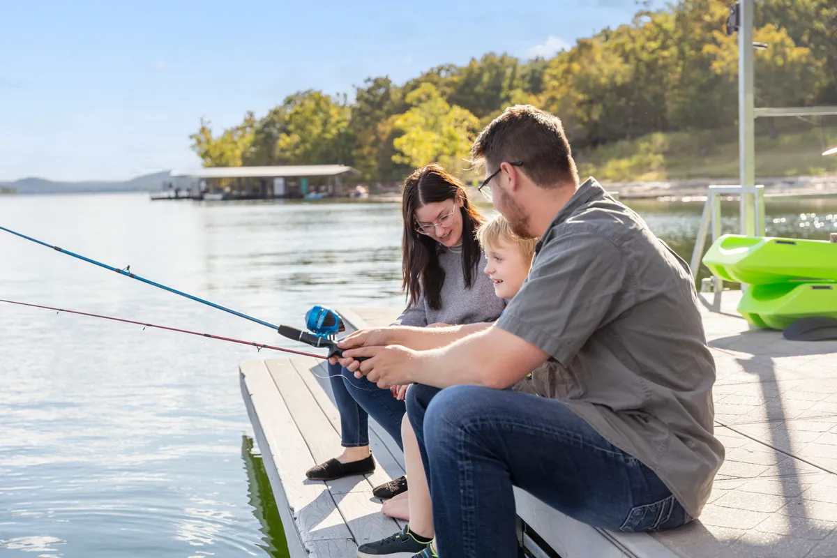 A smiling family stands on the waterfront, casting fishing rods into the calm lake. The scene is set with lush greenery and sparkling water, and the family appears to be sharing laughter and stories as they fish together. The atmosphere is happy and serene, capturing a classic lakeside bonding moment.