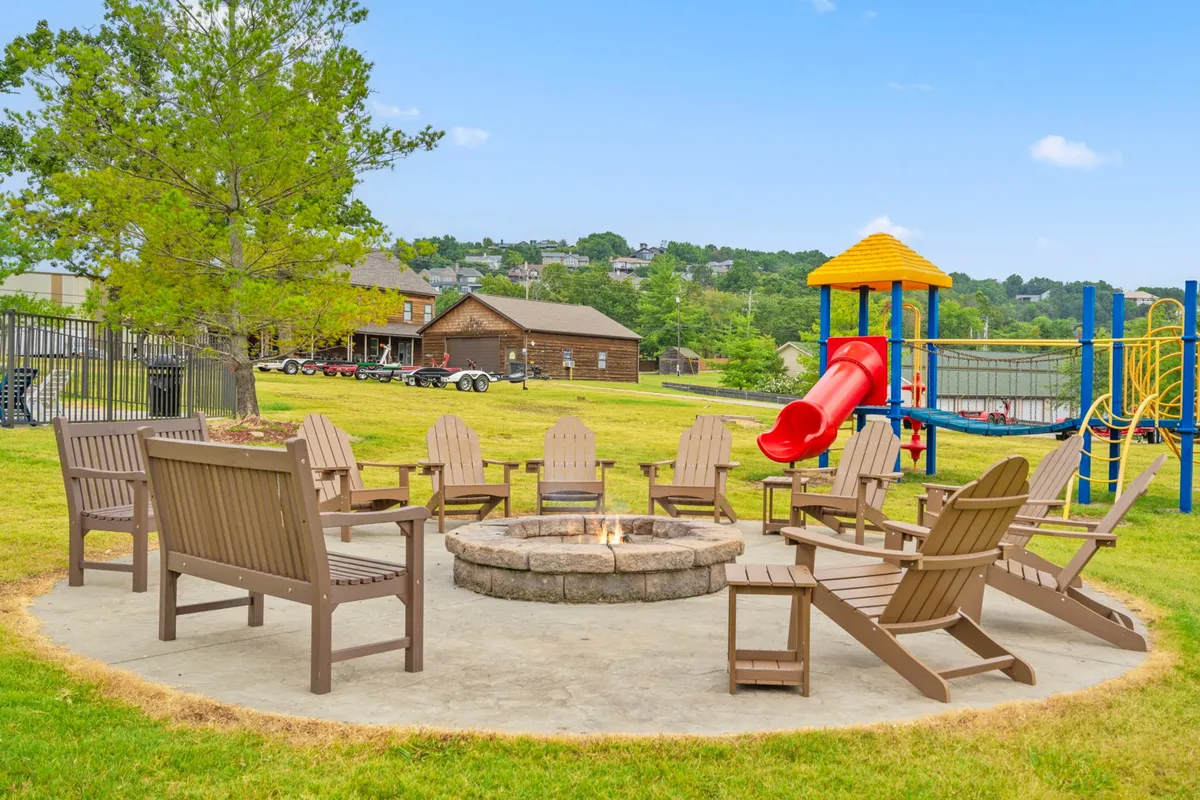 A cozy fire pit surrounded by Adirondack chairs and a green lawn, with playground equipment visible in the background. The sky is blue and clear, and the setup invites campfire gatherings and storytelling as dusk falls.
