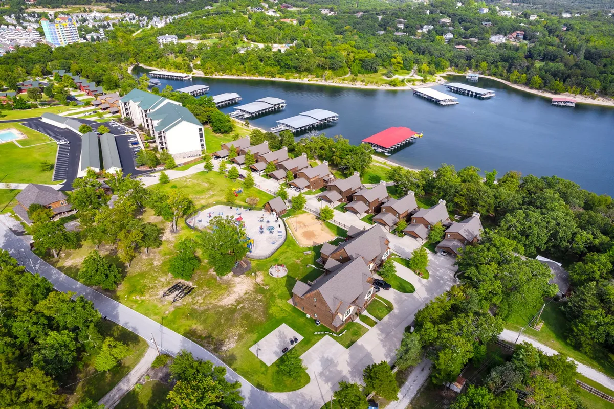 A wide-angle exterior image capturing the lake, docked boats, a green-roofed building, and the playground area surrounded by trees. The image evokes a sense of lakeside community, with children playing and families gathering near the water.