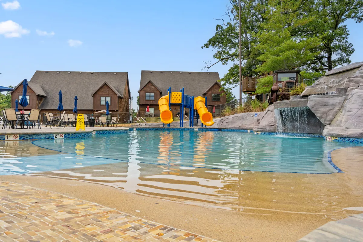 A lively outdoor scene featuring the swimming pool with a water slide, surrounded by cabins and a cascading waterfall. Children and families are seen enjoying the sun, playing in the pool, and relaxing poolside.
